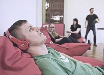 A young Salsita employee lying on a pink bean bag with red headphones while colleagues work in the background