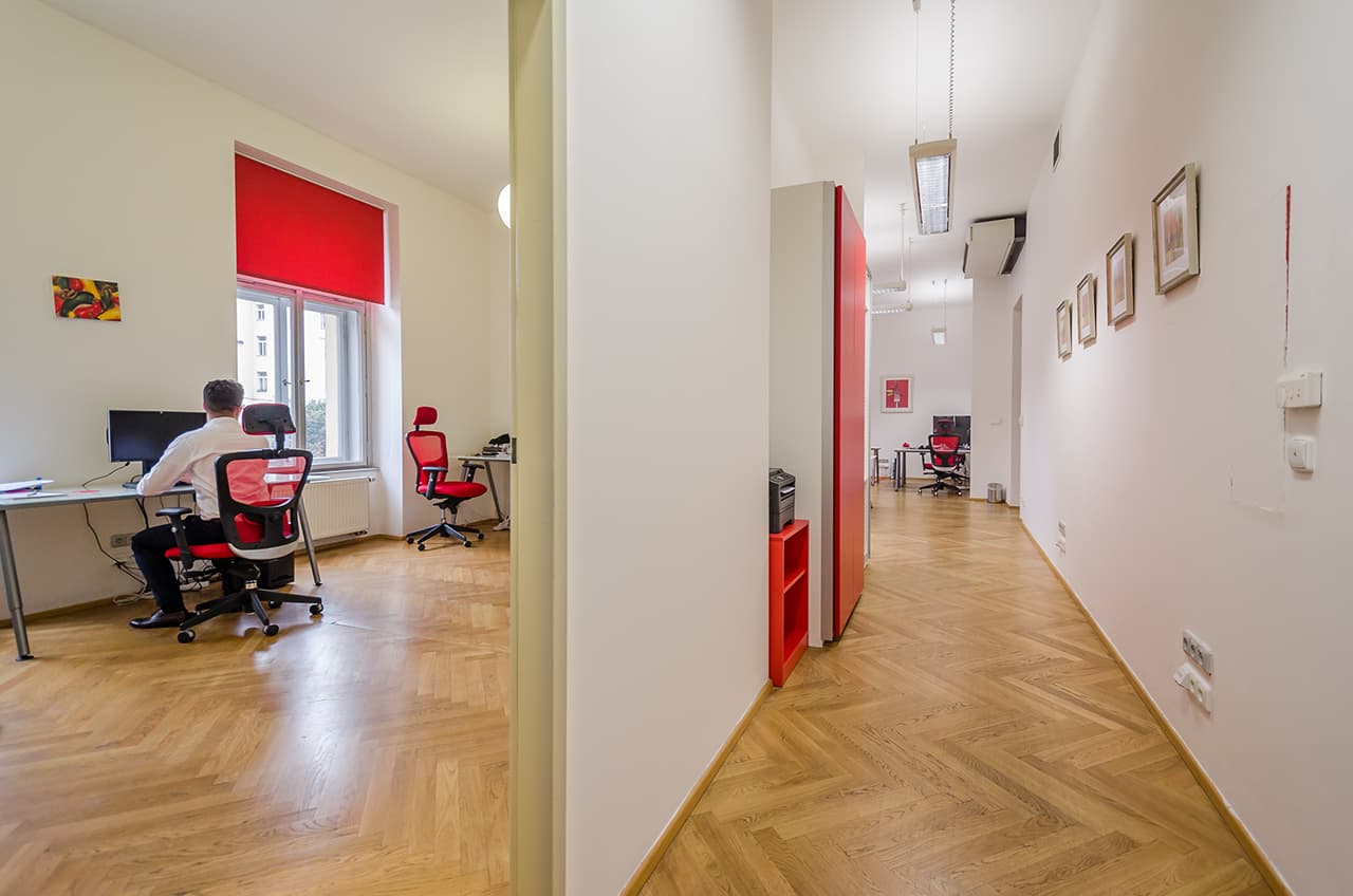 Salsita office space with wooden flooring, white walls, red accents and rows of desks – one person working at a window lit by a red window shade